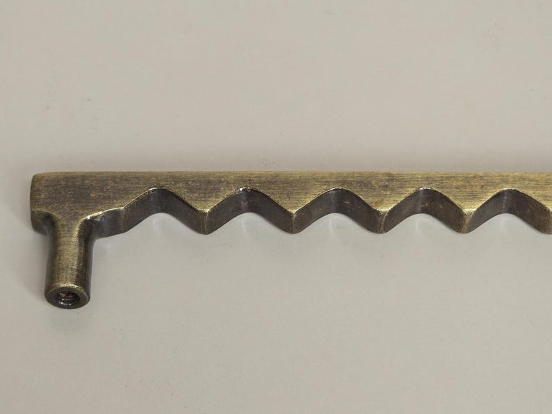 May include: A bronze-colored cabinet pull with a unique, wavy design. The hardware features a series of zig-zag patterns along its length, with a small, cylindrical projection on one end for mounting. This decorative pull adds a vintage touch to furniture.