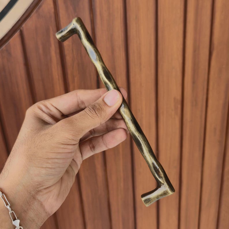 May include: A bronze-colored cabinet pull with a rustic, branch-like design. The handle is held against a brown wooden background. The pull is approximately 10 inches long.