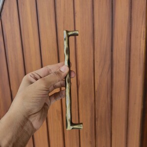 May include: A bronze-colored door handle with a textured, organic design. The handle is held against a wooden door, showcasing its length and shape. The handle's design features a unique, irregular surface.