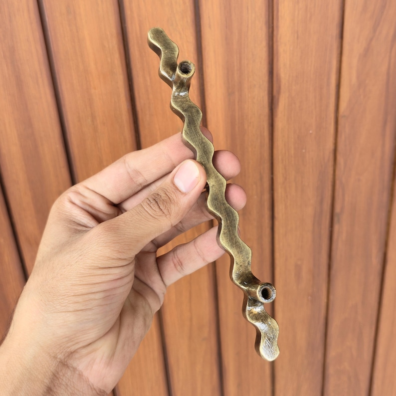 May include: A wavy, antique brass-colored cabinet pull. The decorative handle has a unique, organic shape with two mounting holes. The pull is held against a wooden background.