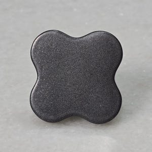 Könnte beinhalten: Ein dunkelgrauer Schrankknopf mit vier Lappen. Die Hardware hat eine strukturierte Oberfläche und eine abgerundete Form. Dieser dekorative Knopf eignet sich für Schubladen, Schränke oder Möbel. Der Knopf ist etwa 3,8 cm breit.