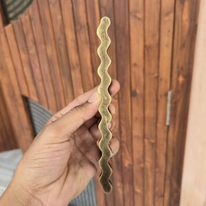 May include: A wavy, brass-colored cabinet pull handle. The handle has a textured surface and a unique, undulating design. The hardware is held against a wooden door with vertical planks.