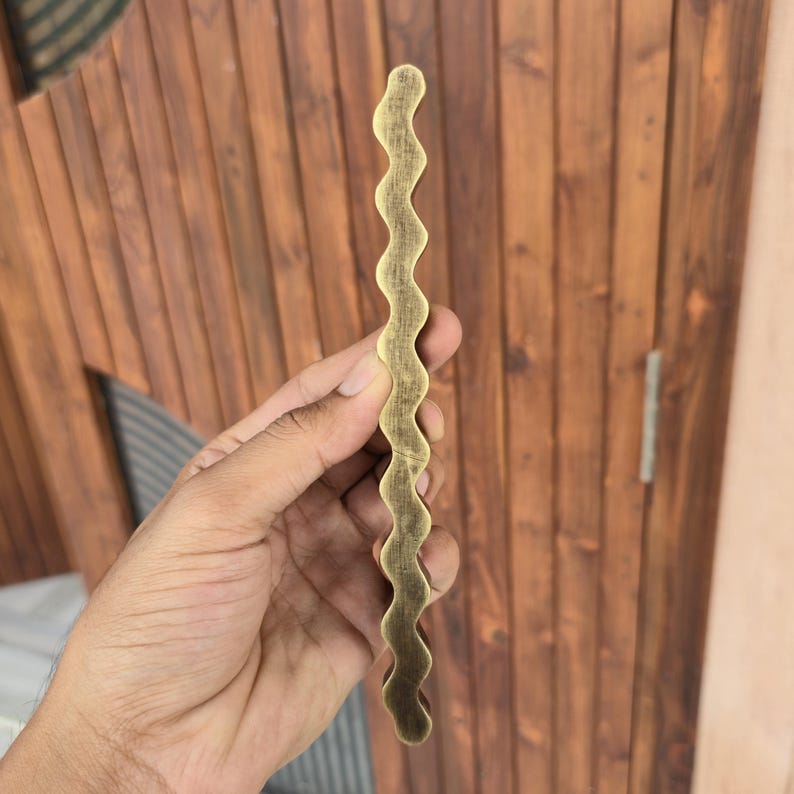 May include: A wavy, brass-colored cabinet pull handle. The handle has a textured surface and a unique, undulating design. The hardware is held against a wooden door with vertical planks.