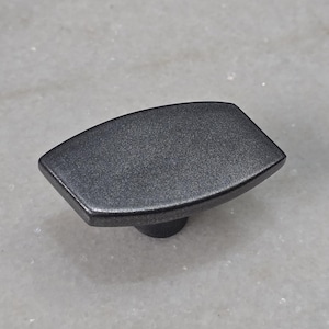 Könnte beinhalten: Ein dunkelgrauer Schrankknopf mit strukturierter Oberfläche. Der Knopf hat eine rechteckige Form mit abgerundeten Kanten und einem kleinen, runden Sockel. Geeignet für Schubladen und Schränke.