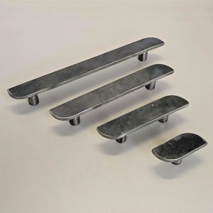 Puede incluir: Cuatro tiradores de armario de metal gris oscuro de diferentes tamaños. Cada tirador tiene forma de barra rectangular con extremos redondeados y un poste cilíndrico para el montaje. Los tiradores están dispuestos sobre un fondo claro.
