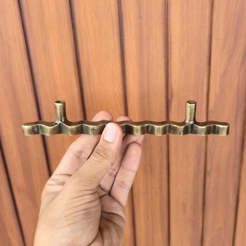 May include: A bronze-colored cabinet pull with a wavy, textured design. The pull has two vertical posts for mounting and a horizontal bar with a series of rounded scallops. The hardware is held against a wooden background.