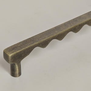 May include: A bronze-toned cabinet pull with a rectangular bar and a series of wave-like cutouts along one edge. The pull has a cylindrical base for mounting. Hardware for furniture.