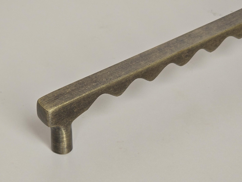 May include: A bronze-toned cabinet pull with a rectangular bar and a series of wave-like cutouts along one edge. The pull has a cylindrical base for mounting. Hardware for furniture.