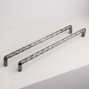 May include: Two silver-toned cabinet pulls with a hammered texture. The pulls are straight with curved ends, designed for easy gripping. They are displayed on a white surface, showcasing their metallic finish and design.