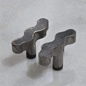 May include: Two metal, wavy-shaped components with a textured, gray finish. Each piece has a short, cylindrical base. The objects are likely hardware or mechanical parts.