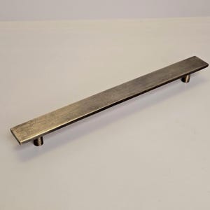 Könnte beinhalten: Ein rechteckiger, bronzefarbener Schrankgriff mit gebürsteter Oberfläche. Die Hardware verfügt über einen flachen, länglichen Stab mit abgerundeten Enden und zwei zylindrischen Pfosten zur Montage. Dieser moderne Griff ist für Schubladen und Schränke geeignet.