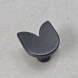 Könnte beinhalten: Ein schwarzer Schrankknopf mit einer einzigartigen, geschwungenen Form. Der Knopf hat eine matte Oberfläche und einen kleinen, runden Sockel. Diese Hardware ist für Schubladen, Schränke oder Möbel geeignet.