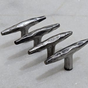 Puede incluir: Cuatro tiradores de armario de metal plateado con textura martillada. Cada tirador tiene un diseño en forma de T, con un asa redondeada y una base cilíndrica corta. Los tiradores están dispuestos en fila, ligeramente inclinados.