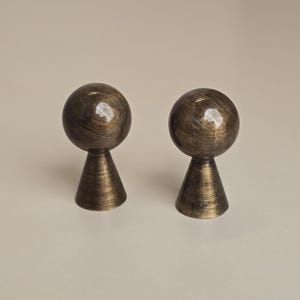 Op de afbeelding: Twee antieke bronzen kastknoppen. Elke knop heeft een bolvormige bovenkant en een conische basis. De knoppen hebben een verweerde afwerking, waardoor ze een vintage uitstraling hebben. Deze decoratieve knoppen zijn geschikt voor meubelrestauratie.