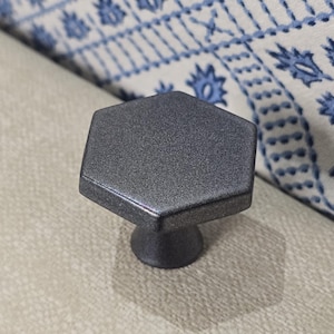 Könnte beinhalten: Ein dunkelgrauer, sechseckiger Schrankknopf mit strukturierter Oberfläche. Der Knopf hat einen kurzen, zylindrischen Sockel. Der Hintergrund umfasst einen Stoff mit weißem und blauem Muster.