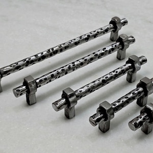 Puede incluir: Juego de cuatro tiradores de armario de metal martillado en varios tamaños. Los tiradores tienen un acabado gris oscuro texturizado con un patrón martillado. Cada tirador tiene una base rectangular y un tirador cilíndrico.