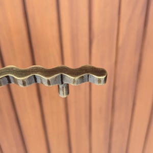 May include: Close-up of a decorative bronze-colored cabinet pull with a wavy design. The pull has a small, cylindrical projection extending from the bottom. The background is a series of vertical, reddish-brown wooden planks.