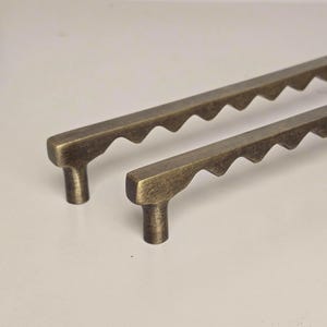 Op de afbeelding: Twee antieke messing kastgrepen met een uniek geschulpte randontwerp. De grepen hebben een rechthoekige basis en een T-vormige handgreep. De messing afwerking geeft ze een vintage uitstraling, perfect voor meubels.