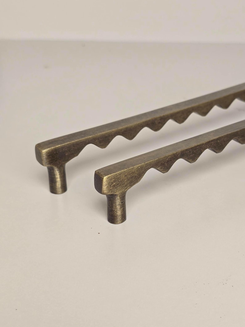 May include: Two antique brass cabinet pulls with a unique scalloped edge design. The pulls have a rectangular base and a T-shaped handle. The brass finish gives them a vintage look, perfect for adding a touch of elegance to furniture.