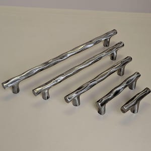 Peut inclure: Un ensemble de six poignées de meubles en métal argenté de différentes longueurs. Chaque poignée présente un design texturé et organique, ressemblant à une branche d'arbre. Les poignées sont disposées en diagonale sur un fond neutre.