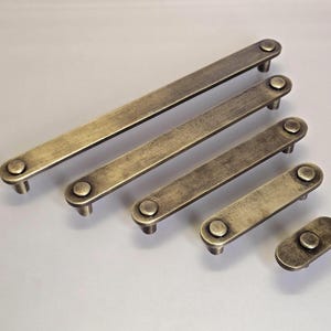 May include: A set of antique-style cabinet handles in a brassy finish. The handles are rectangular with rounded ends and circular knobs. The sizes vary, with a distressed, aged look. Ideal for furniture restoration or adding a vintage touch.