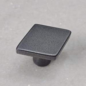 Puede incluir: Un tirador de armario cuadrado de color gris oscuro con un acabado texturizado. El tirador tiene forma de diamante y una base cilíndrica corta. Adecuado para cajones y armarios.
