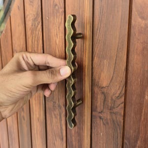 May include: A wavy, antique brass door pull handle. The handle is held against a wooden door with vertical planks. The handle has a unique, organic shape and is mounted with two small, round posts.