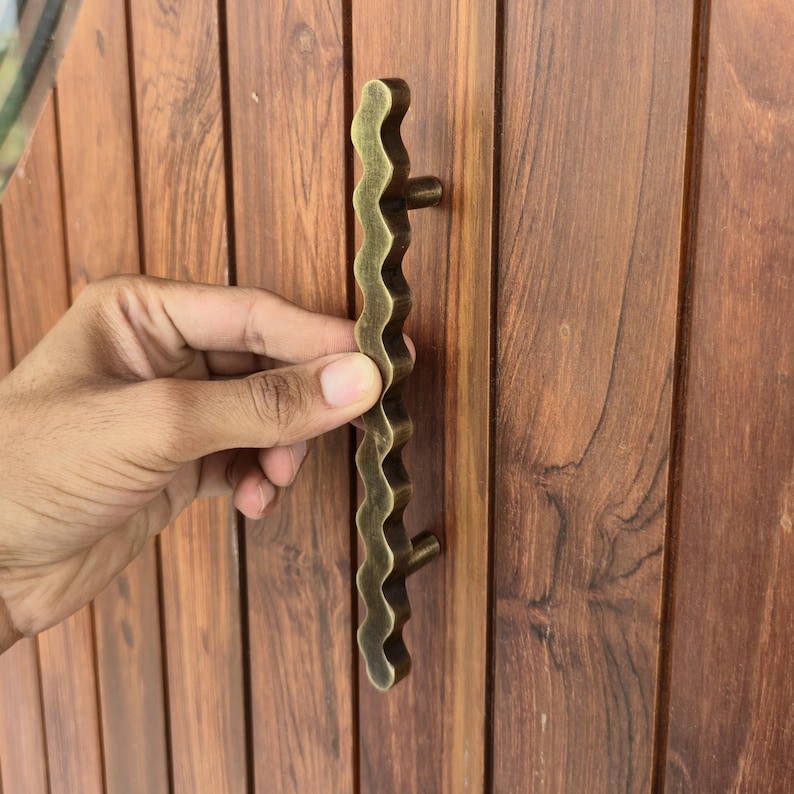 May include: A wavy, antique brass door pull handle. The handle is held against a wooden door with vertical planks. The handle has a unique, organic shape and is mounted with two small, round posts.