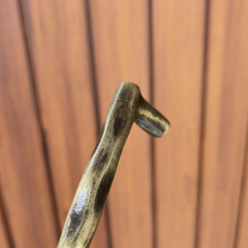 May include: A wooden handle with a curved design and a small, perpendicular extension at the top. The wood has a dark, stained finish with lighter streaks, creating a textured appearance. The handle is angled, suggesting it is part of a larger tool or object.