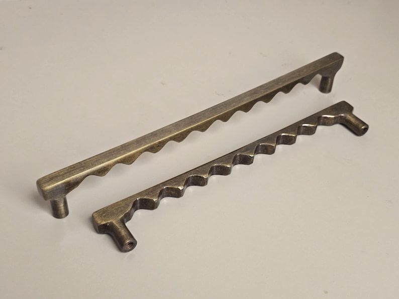 May include: Two antique bronze-colored cabinet pulls. The pulls have a unique design with a series of wave-like cutouts along the top edge. One pull is approximately 10 inches long, and the other is about 6 inches long.