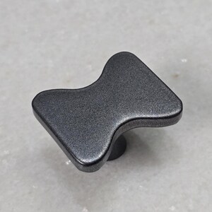 Könnte beinhalten: Ein dunkelgrauer Schrankknauf mit einem einzigartigen, eckigen Design. Der Knauf hat eine strukturierte Oberfläche und einen kleinen, runden Sockel. Geeignet für Schubladen und Schränke.