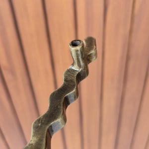 May include: Close-up of a bronze-colored, wavy metal object. The object has a textured surface and a cylindrical opening at the top. The design is reminiscent of a stylized, abstract form, possibly a decorative element or handle.