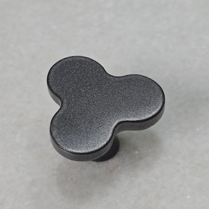 Könnte beinhalten: Ein dunkelgrauer Schrankknauf mit einem dreilappigen Design. Der Knauf hat eine strukturierte Oberfläche und einen kleinen, runden Sockel. Diese dekorative Hardware ist für Schubladen und Schränke geeignet.