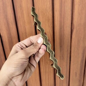 May include: A wavy, antique brass-colored cabinet pull handle. The handle has a unique, organic shape and is held against a wooden background. This decorative hardware is suitable for drawers and cabinets.