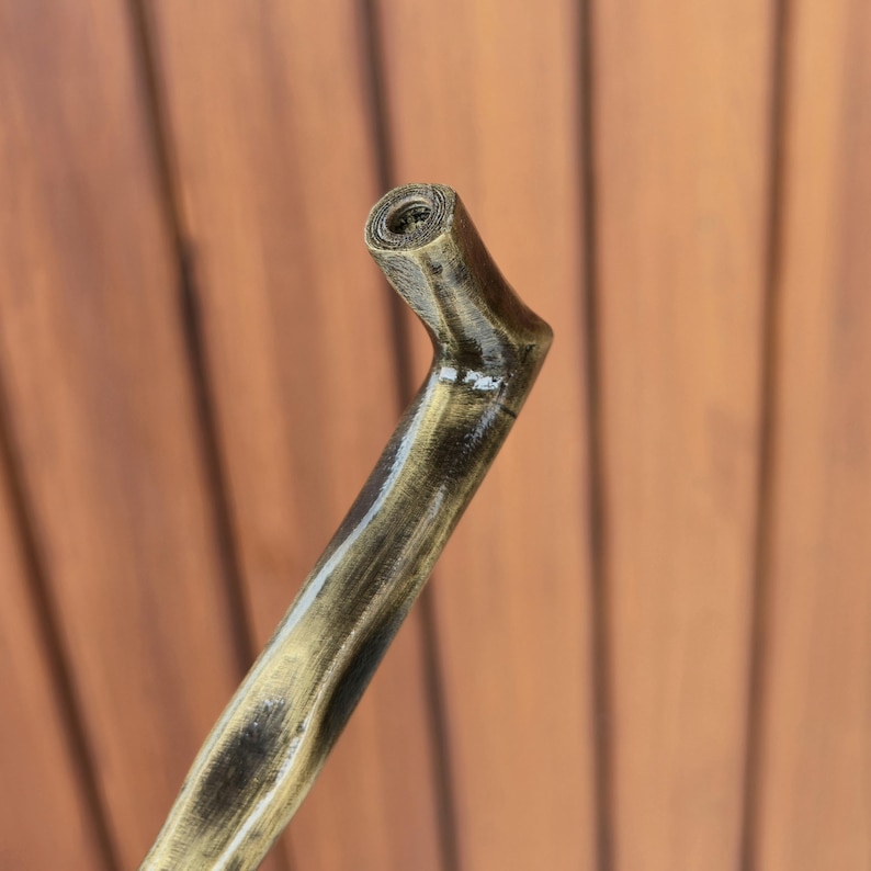 May include: A wooden walking cane with a curved handle. The cane is painted with a gold and black finish, giving it a rustic appearance. The handle is rounded and has a hole at the top.