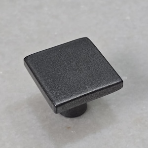 Könnte beinhalten: Quadratischer, mattschwarzer Schrankknopf mit strukturierter Oberfläche. Der Knopf hat einen kurzen, runden Sockel. Die Hardware befindet sich vor einem hellgrauen Hintergrund. Geeignet für Schubladen und Schränke.