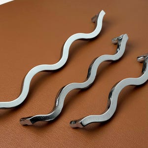 May include: Three silver-colored metal cabinet handles with a distinct wavy, serpentine shape. Each handle features a smooth, reflective finish and two mounting points at the ends, designed for attachment to furniture or cabinetry doors.