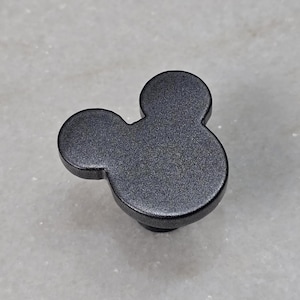 Op de afbeelding: Zwarte kastknop in de vorm van Mickey Mouses hoofd. De knop heeft een getextureerd oppervlak en een kleine, ronde basis. De knop is van metaal en wordt waarschijnlijk voor meubels gebruikt.