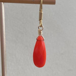 Peut inclure: Une boucle d'oreille unique en forme de goutte avec une pierre précieuse facettée d'un orange-rouge vif, suspendue à un crochet en fil doré. La pierre est fixée par un délicat enroulement de fil doré qui la relie au crochet, créant un design simple.