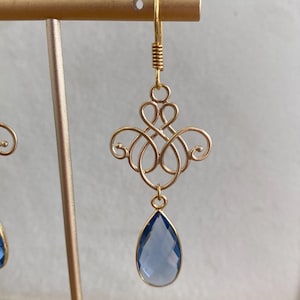 May include: A gold-toned earring featuring a decorative wire-work filigree component with symmetrical scrolls and loops. A faceted, teardrop-shaped blue crystal is suspended from the bottom of the metal design, attached by a small gold-toned jump ring.
