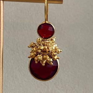 May include: A gold drop earring featuring a small round faceted deep red stone at the top and a larger round faceted deep red stone at the bottom, separated by a cluster of small, textured, metallic gold beads in the center.