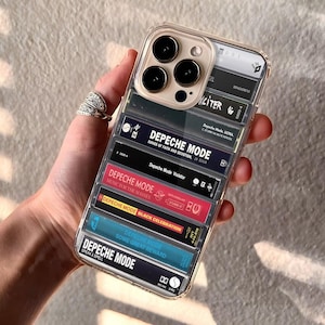 May include: Transparent phone case with a design of stacked cassette tapes. The tapes feature the band name "Depeche Mode" and album titles such as "Violator" and "Some Great Reward". The case has a clear border and a camera aperture.