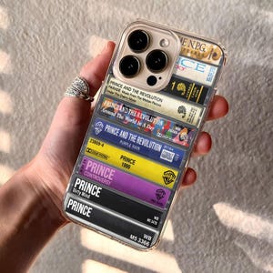 May include: Clear phone case featuring a design of stacked cassette tapes. The tapes display album titles like "Parade," "Around the World in a Day," and "Purple Rain." The case has a clear back and a protective bumper.