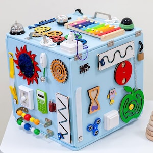 Puede incluir: Un cubo sensorial azul claro con varios elementos interactivos, incluyendo engranajes, un xilófono, una campana y un spinner. El cubo también presenta una manzana de madera y un juguete apilable de madera. El juguete está diseñado para el desarrollo infantil temprano.