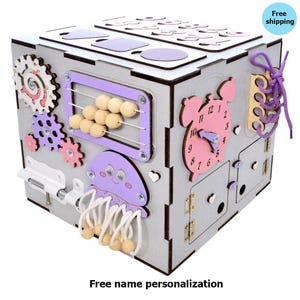 May include: A gray wooden activity cube with various interactive elements. Features include gears, an abacus, a clock, and a jellyfish design. The cube has a variety of colors, including pink, purple, and natural wood tones. The text "Free name personalization" is visible.