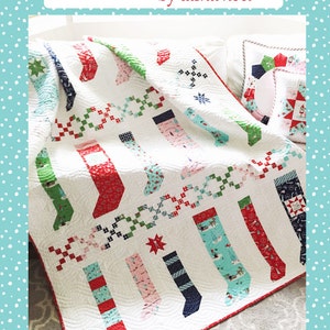 May include: A white quilt with a pattern of colourful stockings. The quilt is folded and displayed on a white sofa. The quilt is titled "Pixie Stockings by Tasha Noel" and has the number "#506" on the bottom right corner.