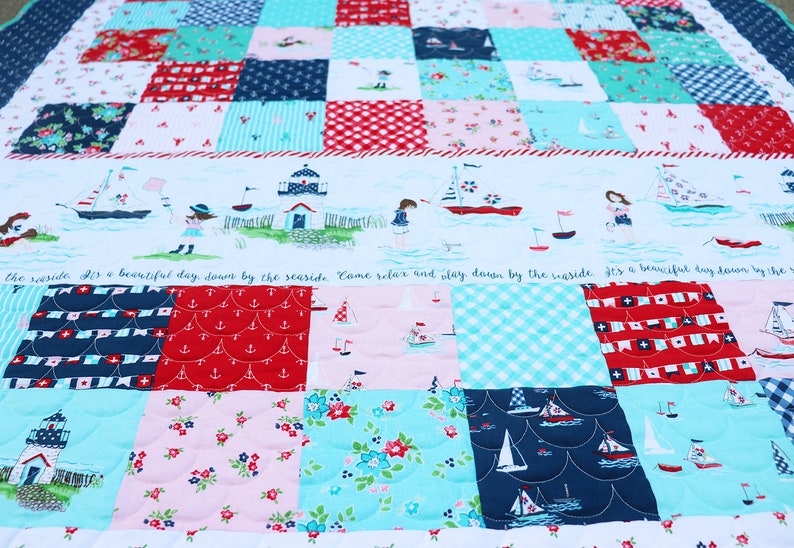 Pode incluir: Um cobertor de retalhos com tema n&aacute;utico. O cobertor &eacute; feito de v&aacute;rios tecidos em tons de azul, vermelho, branco e rosa. O cobertor apresenta uma variedade de padr&otilde;es, incluindo &acirc;ncoras, veleiros e conchas. O cobertor &eacute; guarnecido com uma bainha branca. O texto no cobertor diz: "&Eacute; um dia lindo &agrave; beira-mar. Venha relaxar e brincar &agrave; beira-mar."
