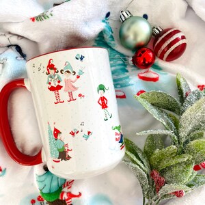 May include: White ceramic mug with a red rim and handle. The mug features a festive design of elves singing carols and a Christmas tree. The mug is sitting on a white and blue patterned blanket with red and green Christmas ornaments.