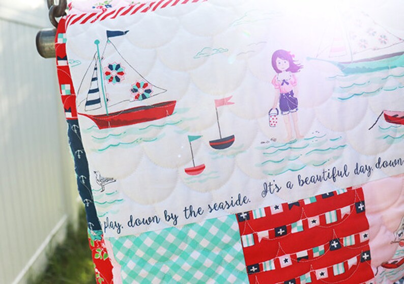 Pode incluir: Um edredom de retalhos branco e azul com tema n&aacute;utico. O edredom apresenta veleiros, bandeiras e uma menina segurando um balde. O texto "It's a beautiful day down by the seaside." est&aacute; costurado no edredom.