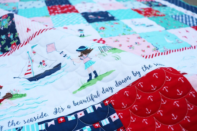 Pode incluir: Uma colcha branca com um veleiro azul, uma menina com uma pipa e o texto "It's a beautiful day, down by the sea" em um fundo branco. A colcha &eacute; feita de quadrados de patchwork com diferentes padr&otilde;es, incluindo listras vermelhas e brancas, listras azuis e brancas e um fundo vermelho com &acirc;ncoras brancas.